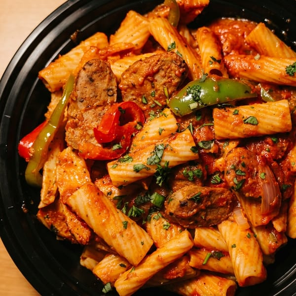 Black bowl of rigatoni pasta with tomato sauce, sausage, peppers, onions, and fresh herbs on a wooden table.