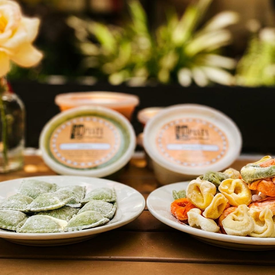 Two plates of colorful fresh pasta, including green ravioli and assorted tortellini, sit on a sunlit wooden table beside a flower.